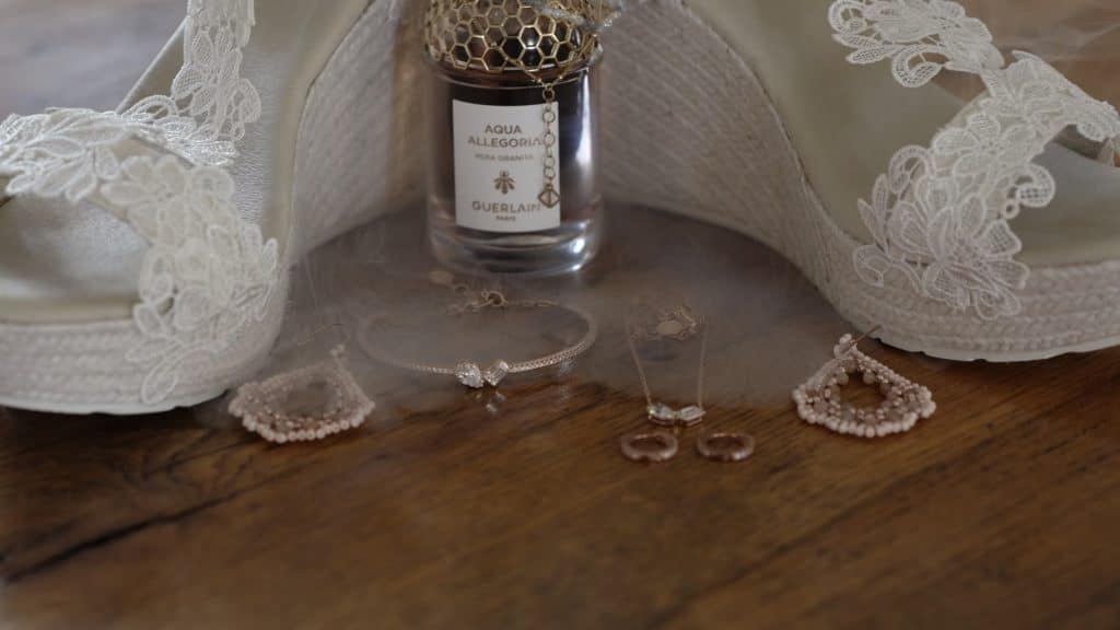 "Ein Stillleben mit Brautschuhen aus Spitze, einer Parfümflasche 'Aqua Allegoria' von Guerlain, und verschiedenen Schmuckstücken, darunter Ohrringe, ein Armband und eine Halskette. Die Szene ist elegant arrangiert und auf einem Holztisch fotografiert,​⬤